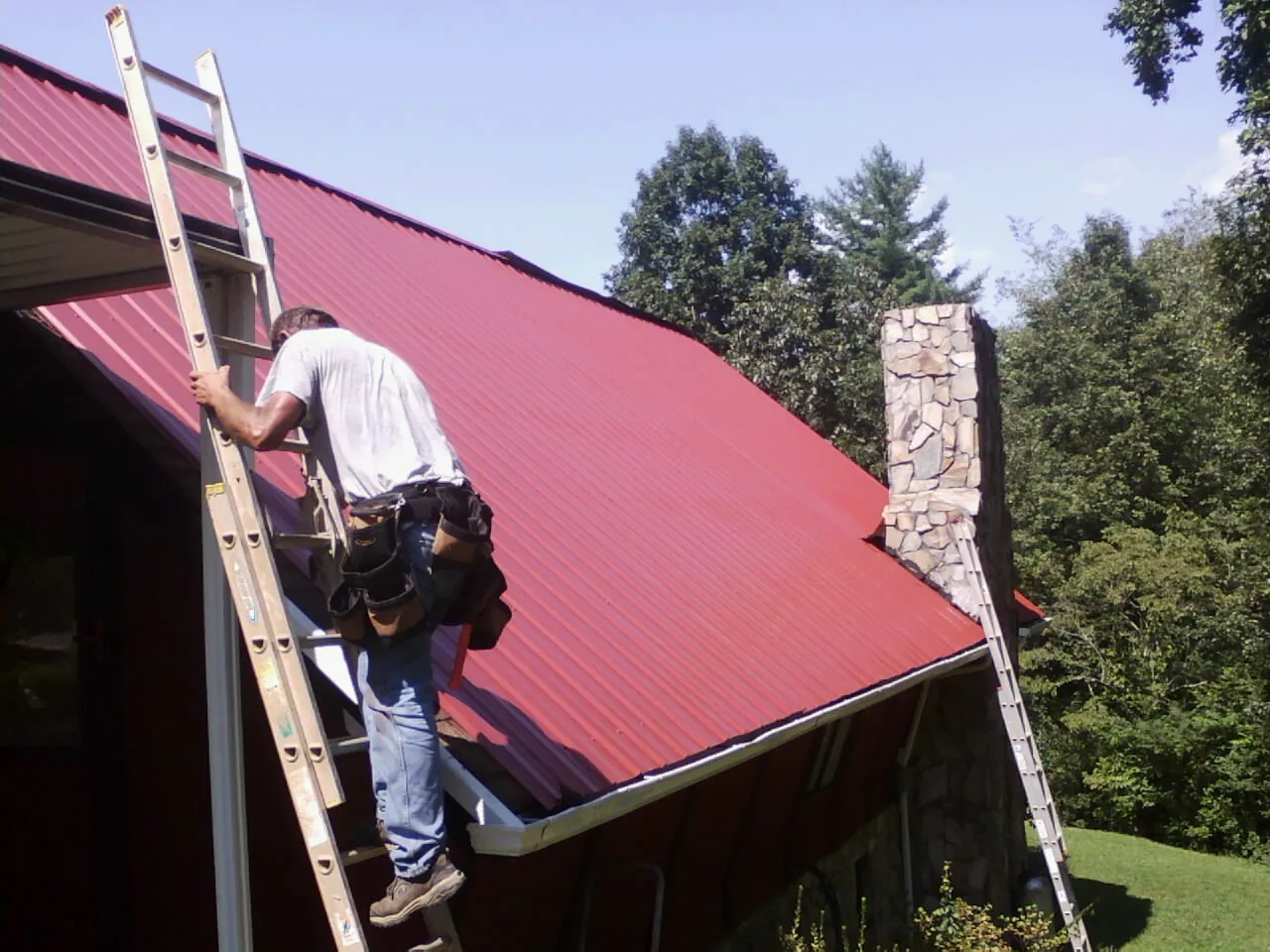 Roofer on a ladder beside a red metal roof and stone chimney — Soffit & Fascia Repair by Roofing Brothers in Montauk